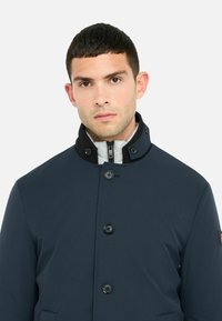 Giacca blu navy con colletto alto, bottoni frontali e fodera interna a contrasto. Tessuto liscio e strutturato con tasche laterali.