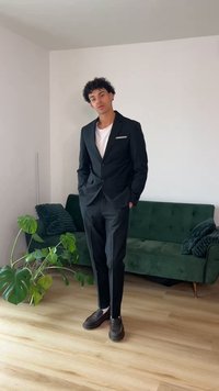 Göndör hajú fiatal férfi fekete öltönyben és fehér ingben, egy olyan szobában áll, ahol zöld kanapé és nagy levelű növény található.