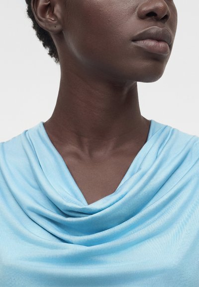 Κοντινό πλάνο ενός ατόμου με σκούρο δέρμα που φοράει ένα ανοιχτό μπλε τοπ με πτυχώσεις, εμφανίζοντας τον λαιμό, τη γωνία της γνάθου και τα χείλη ενάντια σε απλό φόντο.