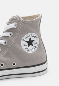 Gray canvas high-top sneaker with white laces, contrasting black rubber sole, and a circular black and white "Converse All Star" logo patch.