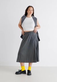 Gonna grigia a pieghe, di media lunghezza, con vita alta. Indossata con un crop top bianco e un gilet grigio. Scarpe nere e calzini gialli brillanti.