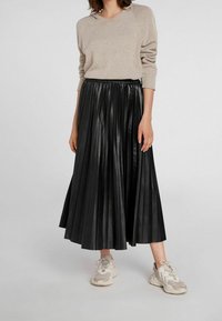 Zwarte plissee rok van leer met een elastische tailleband. Gepaard met een lichtbeige sweater en lichte sneakers.