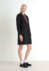 Čierne sako šaty s priliehavým strihom, dvoma prednými vreckami a jedným gombíkom, skombinované s tmavým topom a lesklými čiernymi mokasínami.