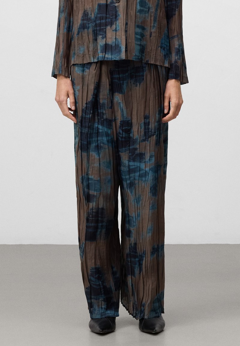 Twee-delige outfit in geplooide stof met een bruine basis en blauwe abstracte patronen. Losvallend, gecombineerd met zwarte schoenen.