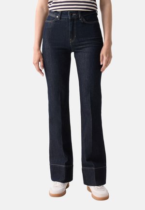 Person iført mørkeblå vidde jeans med opsmøgede kanter, hvide sneakers med brune såler og en sort-hvid stribet top.