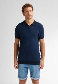 Tmavě modré polo tričko s texturovaným vlnitým vzorem, krátkými rukávy, tříknoflíkovou légou a žebrovanými manžetami a lemem. Nosit s světlými denimovými šortkami.