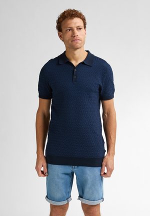 Camisa polo azul marino con patrón de olas texturizado, de manga corta, con tapeta de tres botones y puños y dobladillo de canalé. Combinada con pantalones cortos de mezclilla claros.