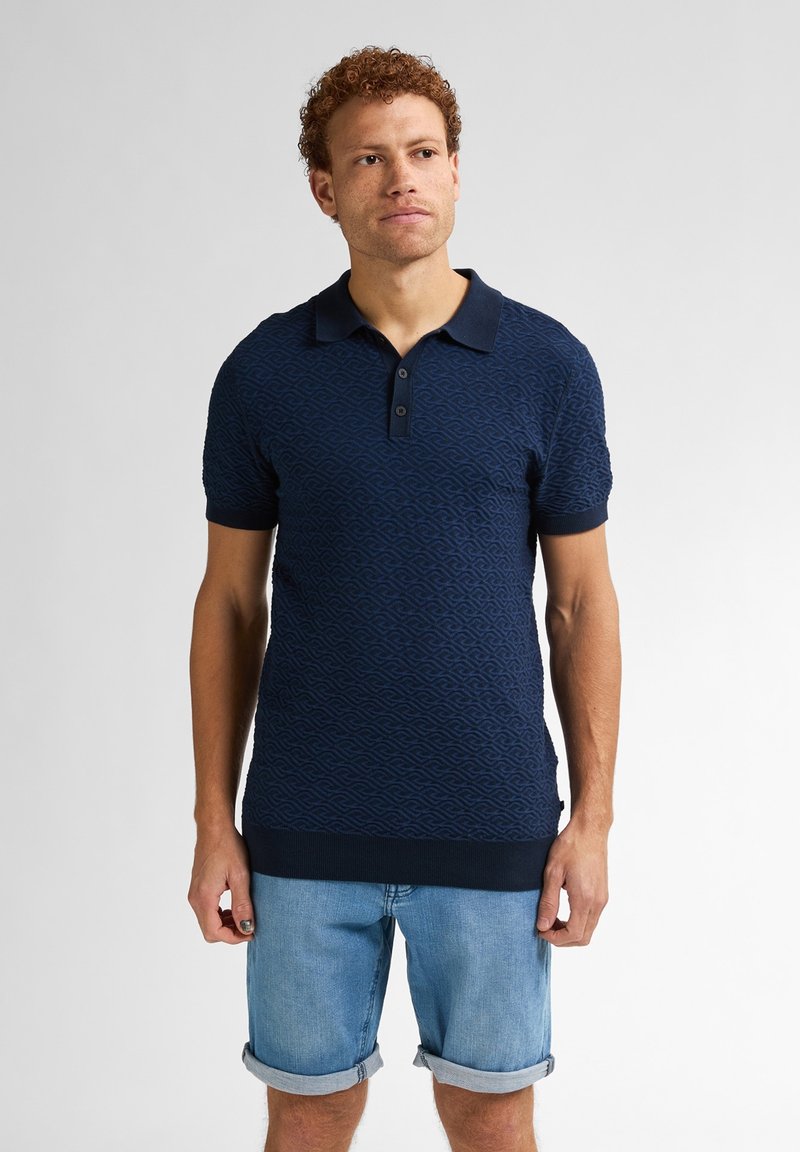 Tmavě modré polo tričko s texturovaným vlnitým vzorem, krátkými rukávy, tříknoflíkovou légou a žebrovanými manžetami a lemem. Nosit s světlými denimovými šortkami.