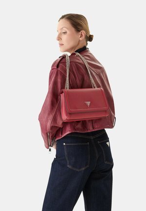 Sac à main en cuir rouge au design structuré, avec une bandoulière en chaîne argentée et un détail logo à l'avant, associé à une veste en cuir bordeaux et un jean noir.