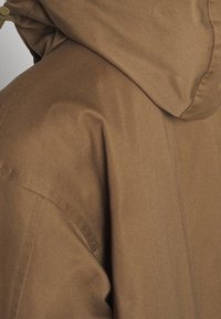 Gros plan d'une veste en tissu marron avec une capuche, montrant les coutures détaillées sur l'épaule et la couture de la capuche.