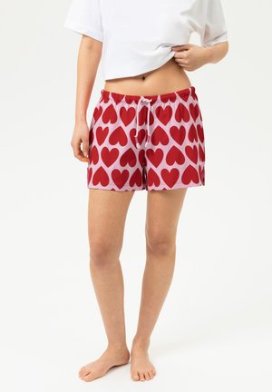 Femme portant un short rose avec un motif de cœur rouge et un t-shirt blanc, debout pieds nus sur un fond clair uni.