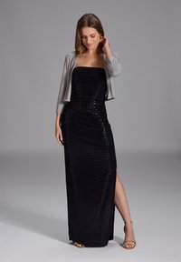 Abito lungo senza spalline in paillettes nere con uno spacco laterale, abbinato a un giubbotto corto argentato. Tacchi dorati con cinghie completano il look. Texture liscia.