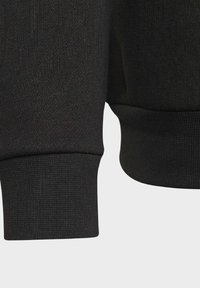 Černá mikina zblízka, s žebrovanými manžetami a lemem. Hladká textura s pleteným vzorem a přiléhavým střihem.