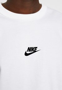 Bílý bavlněný tričko s kulatým výstřihem a černým logem Nike na levé části hrudi, má hladkou texturu a klasický střih.