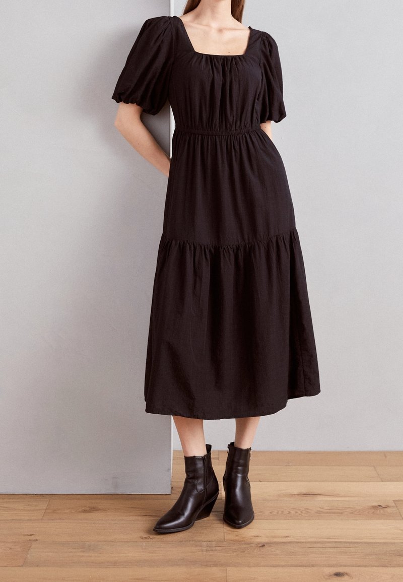 Femme portant une robe midi noire à manches bouffantes et encolure carrée, associée à des bottines noires pointues, debout sur un sol en bois.