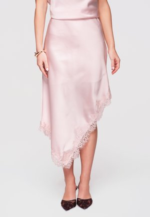 Kobieta w różowej satynowej spódnicy midi z koronkową lamówką, połączona z ciemnymi zdobionymi szpiczastymi szpilkami i złotymi bransoletkami na nadgarstku.