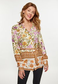 Camicia a fiori bianca, con fiori viola e arancioni, dotata di un nodo in vita e maniche lunghe. Tessuto morbido con colletto.