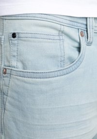 Světle modré džíny z denimu s klasickým designem přední kapsy, viditelným šitím a malým tmavým knoflíkem jako akcentem.