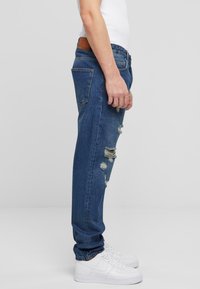 Jeanși din denim albastru cu o croială lejeră, având petice distruse și o etichetă din piele maro închis la talie. Asortați cu adidași albi.