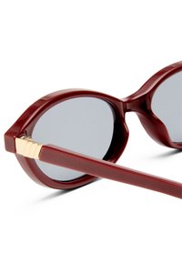 Sunglasses with oversized, oval dark lenses, burgundy plastic frame, and gold metallic accent near the hinge. Smooth texture, modern design.