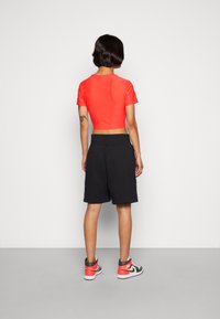 Person med kort hår som bär en röd crop top, svarta knälånga shorts och svarta, vita och röda högsta sneakers, står mot en plain vägg.