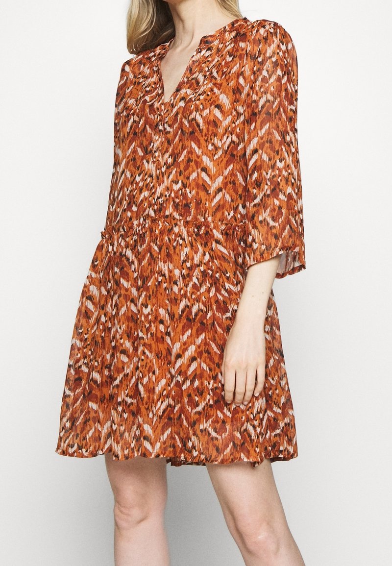 Femme portant une robe orange ample, longueur genou, avec un imprimé animal abstrait et des manches trois-quarts, debout devant un fond uni.