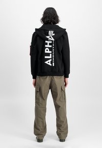 Sort hættetrøje med stort hvidt "ALPHA INDUSTRIES" logo på ryggen, parret med olivengrønne cargo bukser med sidelommer.
