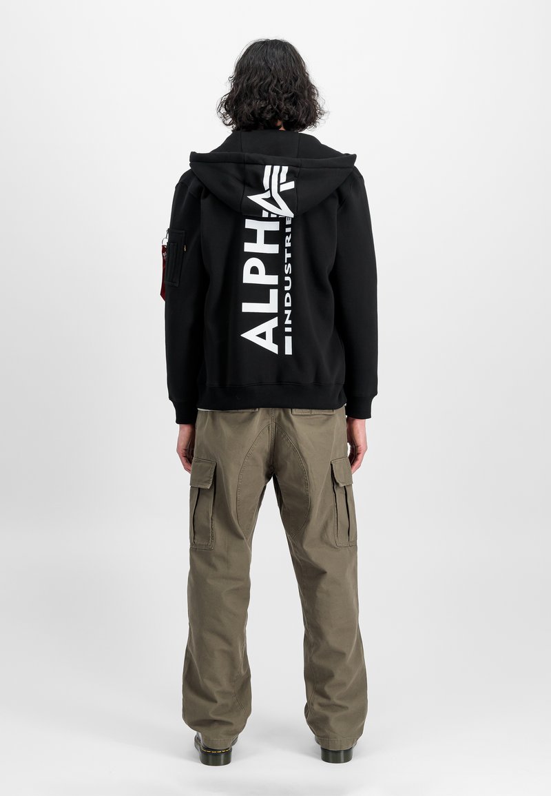 Sort hættetrøje med stort hvidt "ALPHA INDUSTRIES" logo på ryggen, parret med olivengrønne cargo bukser med sidelommer.