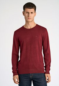Czerwony sweter typu crew-neck wykonany z cienkiej dzianiny, z długimi rękawami i ściągaczami przy mankietach, w połączeniu z niebieskimi dżinsami. Dyskretne logo na piersi.