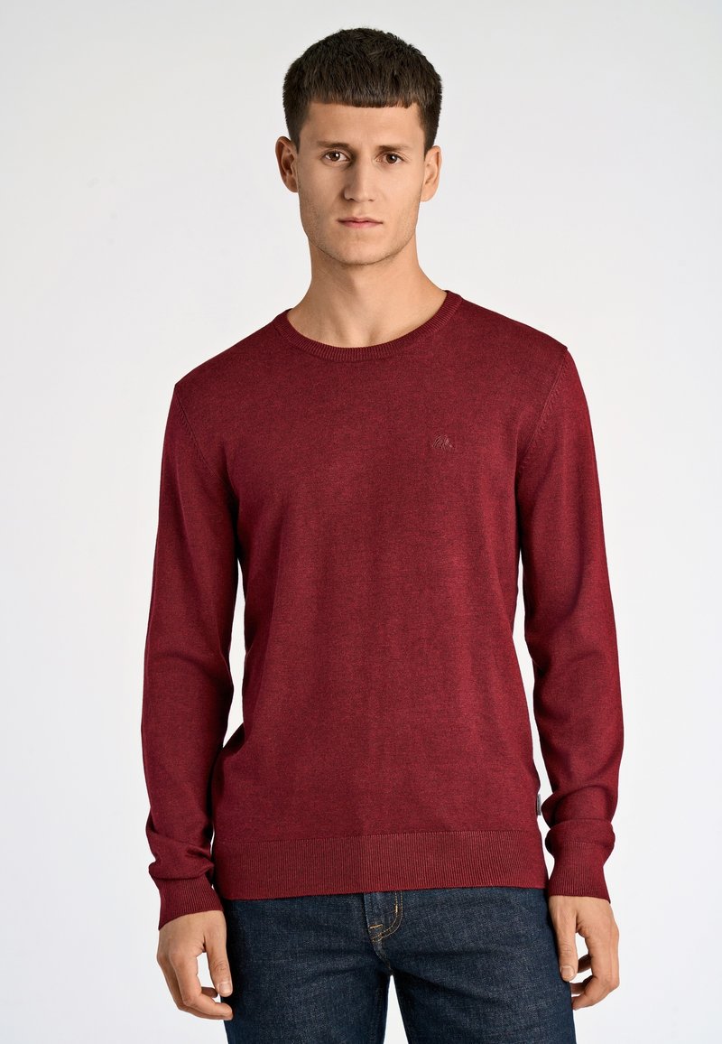 Czerwony sweter typu crew-neck wykonany z cienkiej dzianiny, z długimi rękawami i ściągaczami przy mankietach, w połączeniu z niebieskimi dżinsami. Dyskretne logo na piersi.