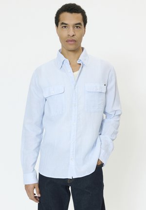 Lyseblå linskjorte med lange ærmer, to brystlommer og knaplukning. Båret over en hvid undertrøje, kombineret med mørke jeans.