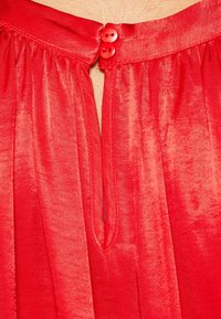 Vêtement rouge avec un design plissé, texture lisse et deux boutons ronds à l'ouverture du cou. Accent sur la couleur vive et les détails du tissu.