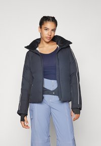 Doudoune noire avec col montant, présentant des rayures blanches sur les côtés et une texture lisse. Associée à un haut bleu marine et un pantalon de ski bleu.