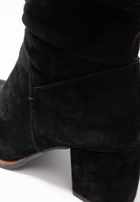 Bottine en daim noir avec un talon carré. Elle présente une finition lisse, des coutures discrètes et un design légèrement froissé.