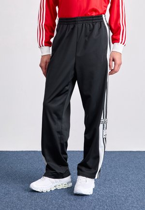 Personne portant un pantalon de survêtement Adidas noir avec des bandes blanches et des baskets blanches debout sur un tapis bleu.