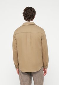 Καφέ χακί πουκάμισο με μακριά μανίκια, μυτερό γιακά, δύο πιέτες στην πλάτη και μανσέτες με κουμπιά· λεία υφή και κομψή εφαρμογή.