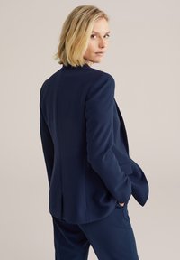 Jūras spēku stilā šūts blazeris no gludas auduma ar pieguļošu siluetu, ar grieztiem uzkabēm un vienu aizmugures venti. Iespējotas priekšējās kabatas un eleganta dizaina.