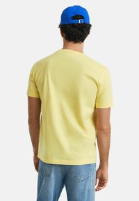 Yellow cotton t-shirt with short sleeves and a round neck, worn with light blue jeans. A blue cap is visible on the person's head.