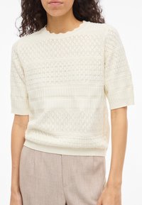Femme portant un top en maille texturé blanc à manches courtes avec motifs en losange et côtelés, associé à un pantalon beige clair.