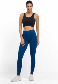 Reggiseno sportivo nero con cucitura frontale, abbinato a leggings a vita alta blu con una texture liscia e dettaglio del logo sulla gamba. Scarpe da ginnastica bianche.