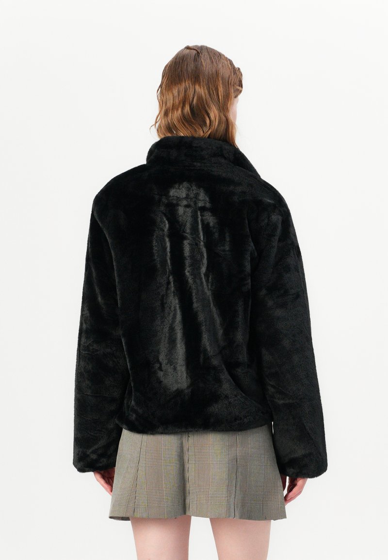 Veste en fausse fourrure noire avec un large col et des manches longues, présentant une texture douce et une coupe ample. Portée par-dessus une jupe grise à motifs.