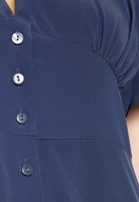 Blusa azul-marinho feita de um tecido leve e suave, com frente abotoada com botões redondos e detalhes franzidos nos ombros.