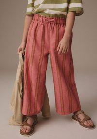 Brede roze linnen broek met verticale rode en oranje strepen, voorzien van een elastische tailleband met een strik, gecombineerd met bruine sandalen.