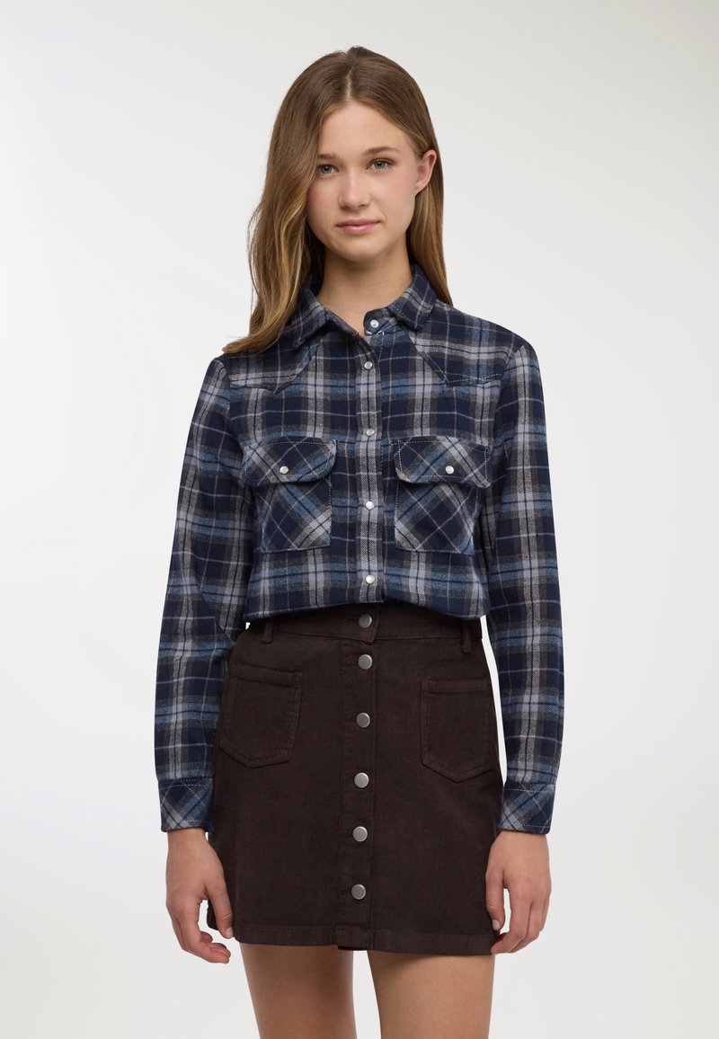 Camicia di flanella a quadri blu con due tasche sul petto, colletto con bottoni e maniche lunghe, abbinata a una mini gonna di velluto a coste marrone con bottoni frontali.