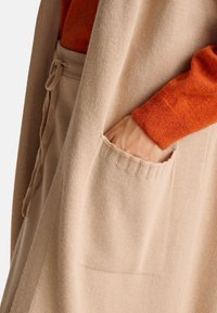 Dlouhý béžový pletený kardigan s texturovaným povrchem, který má dvě boční kapsy a volný střih. Je také viditelná oranžová pletenina.