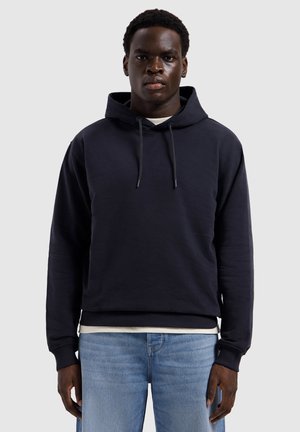 Man draagt een zwarte hoodie en lichtblauwe spijkerbroek, staat rechtop tegen een effen lichtgrijze achtergrond.