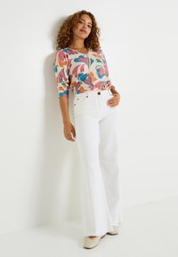 Pull over à motif floral avec des motifs roses, oranges et turquoise ; associé à un jean flare blanc. Tissu texturé et accents de boutons visibles.