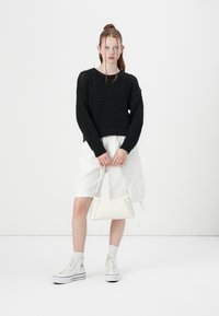 Pull en maille noire avec un motif en dentelle ouverte, shorts cargo blancs et baskets lacées blanches. Tenant un petit sac à main blanc avec des détails en dentelle.