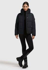 Donna che indossa un giubbotto imbottito nero sopra leggings neri e stivali neri chunky, in piedi con le gambe incrociate su uno sfondo semplice.