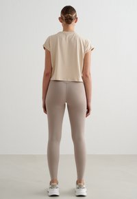Femme avec les cheveux en chignon portant un haut court beige à manches courtes, des leggings taupe et des baskets blanches, debout contre un mur uni.